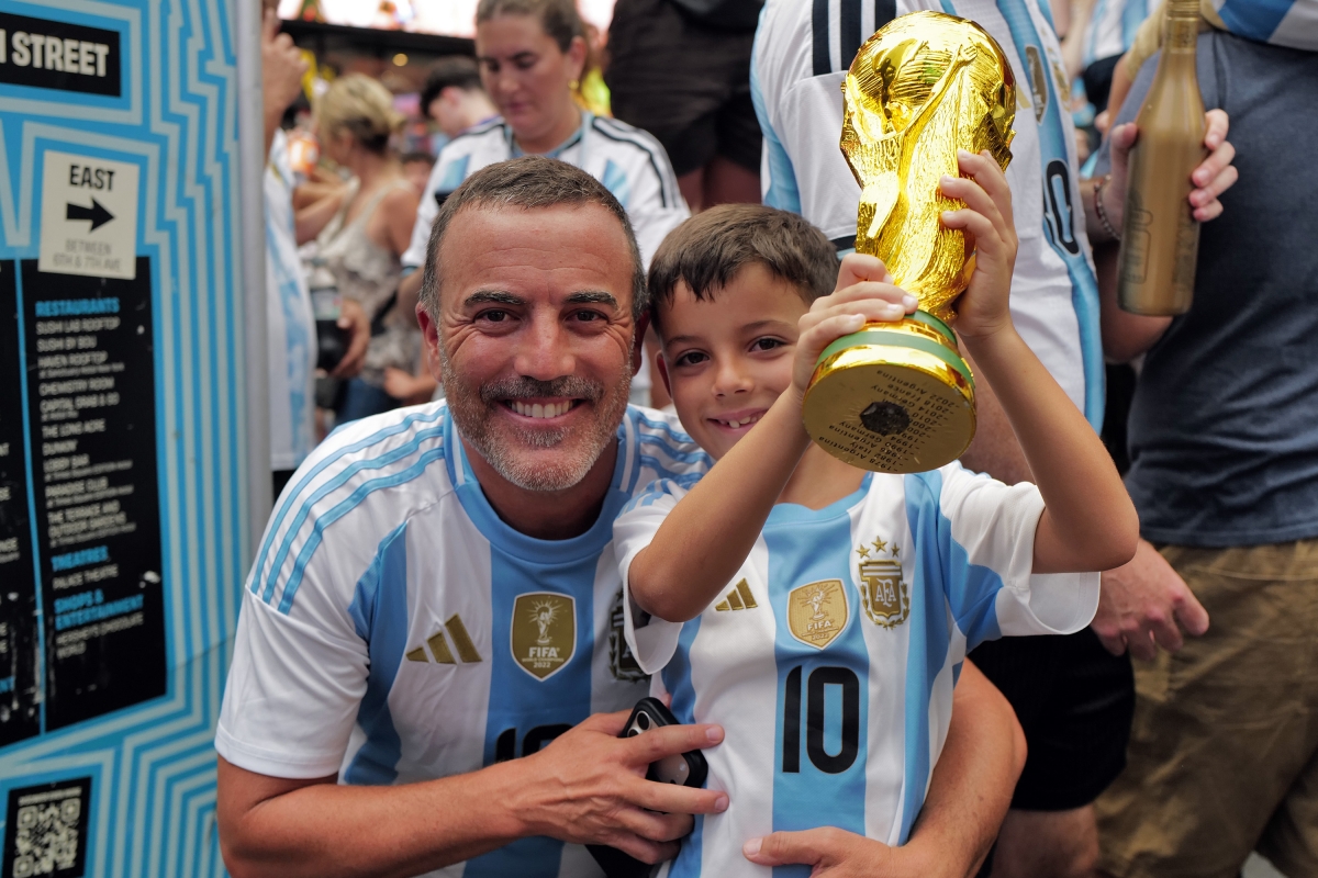 Một em nhỏ trong màu áo Argentina hào hứng nâng mô hình cúp vàng World Cup trước sự cổ vũ của người thân.