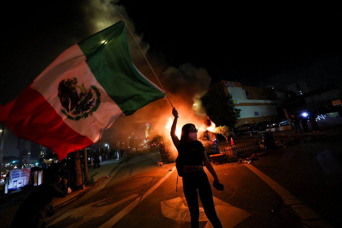 Một người biểu tình vẫy cờ Mexico trước những thùng rác đang cháy trong cuộc biểu tình phản đối chiến dịch truy quét người nhập cư liên bang ở trung tâm thành phố Los Angeles, California, Hoa Kỳ (ảnh: Reuters)